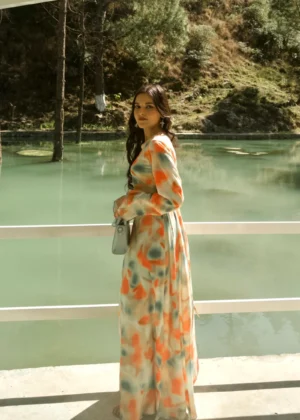 A model in the Coral Breeze Maxi by Rojonera — a vibrant tie and dye maxi dress with soft flared sleeves and a flowy silhouette, photographed lakeside. Crafted from breathable fabric with a lace-up back, this standout piece combines bold color with effortless grace.